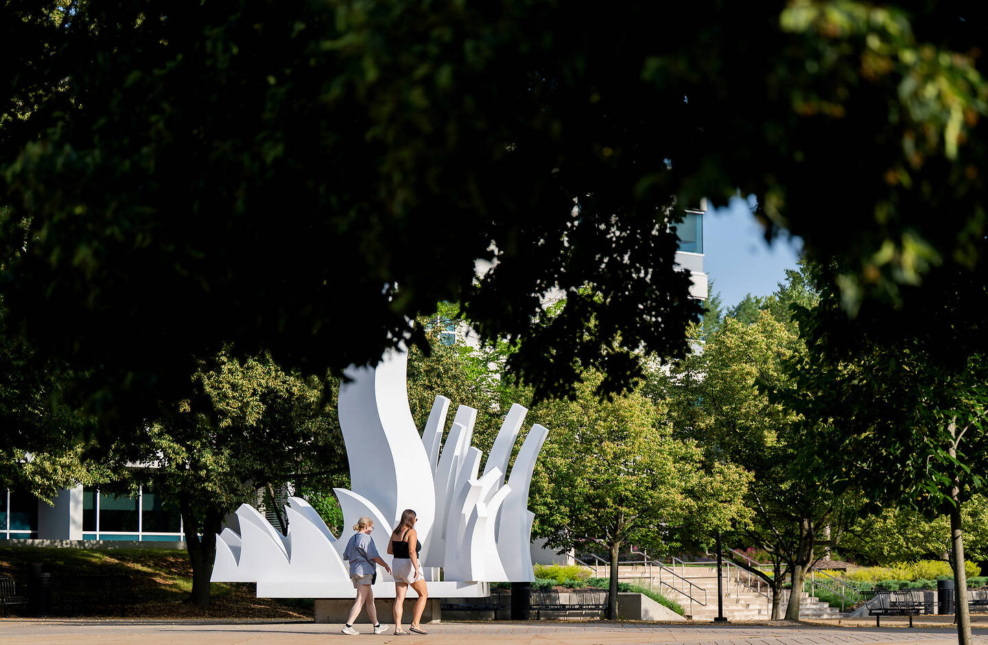 Two individuals walk past a large white abstract sculpture in a park surrounded by trees and modern buildings.